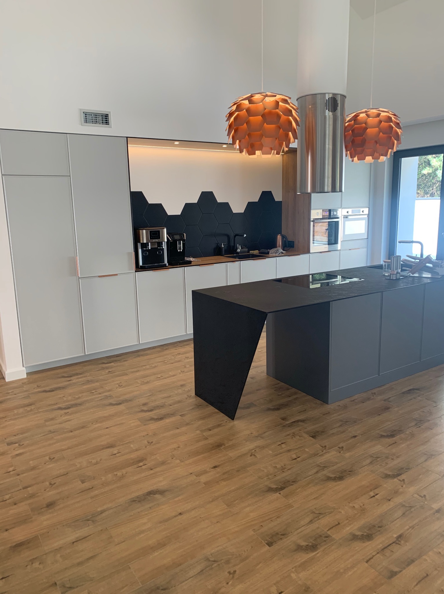 Nowoczesna kuchnia z szarymi szafkami, czarnym blatem wyspy i heksagonalnymi płytkami na ścianie. Nad wyspą wiszą dwie lampy w kształcie kuli i metalowy okap.