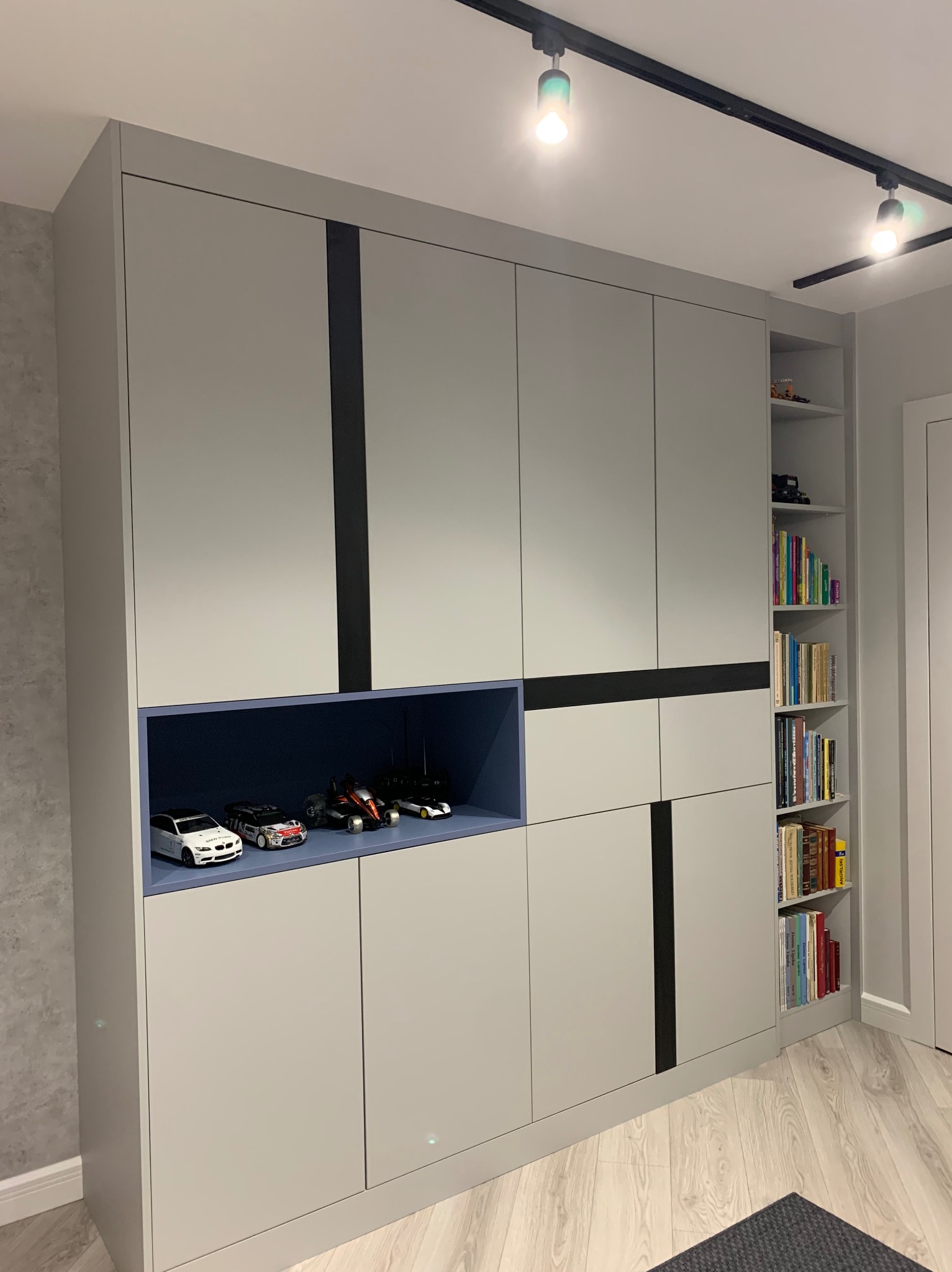 Szara szafa wnękowa z ciemnymi wstawkami i niebieską wnęką, w której stoją modele samochodów. Obok regał z książkami. Oświetlenie sufitowe.