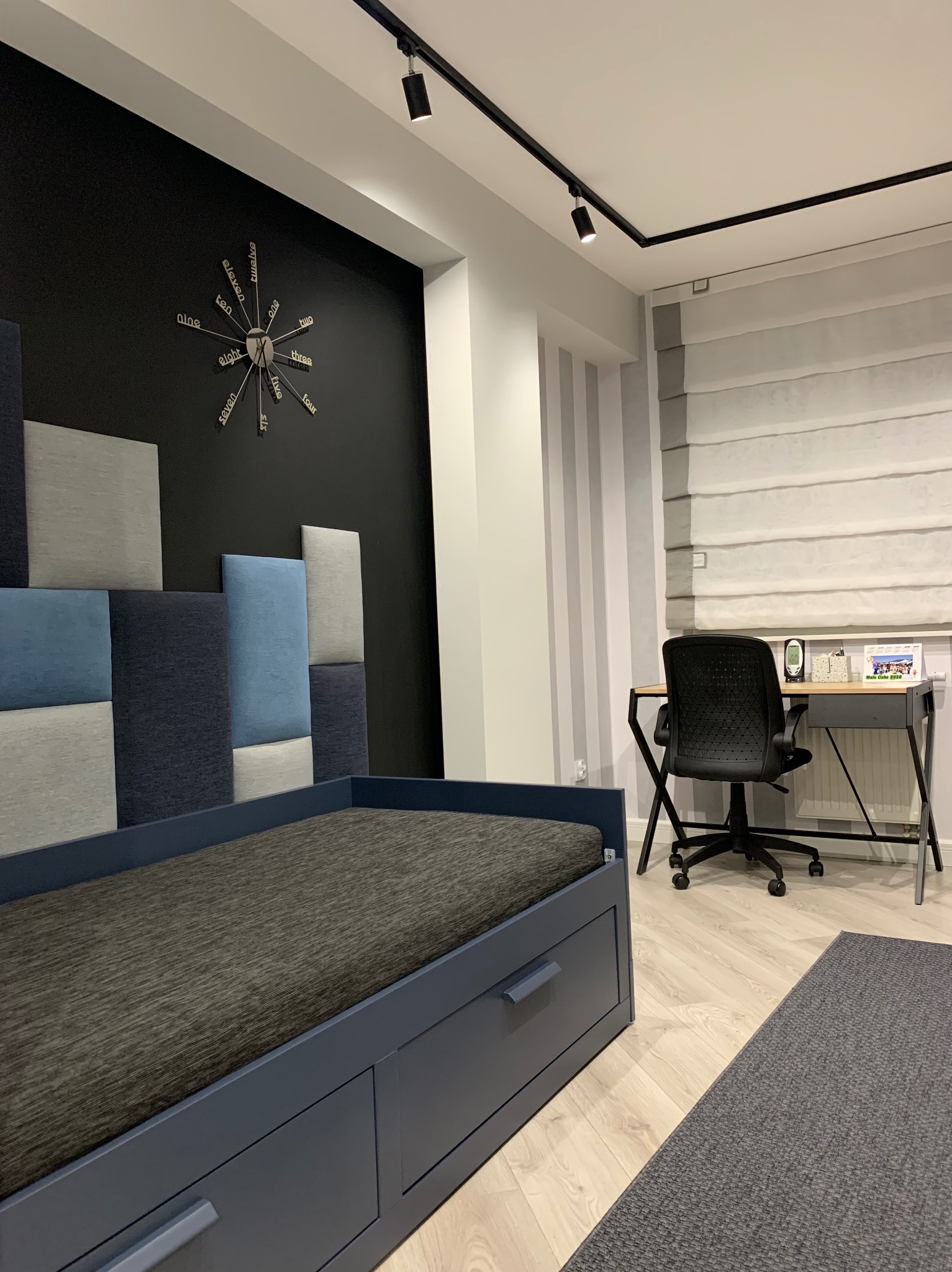 Pokój młodzieżowy z łóżkiem z szufladami i tapicerowanym wezgłowiem w geometryczny wzór. Na ścianie zegar i biurko z krzesłem. Nowoczesne oświetlenie.