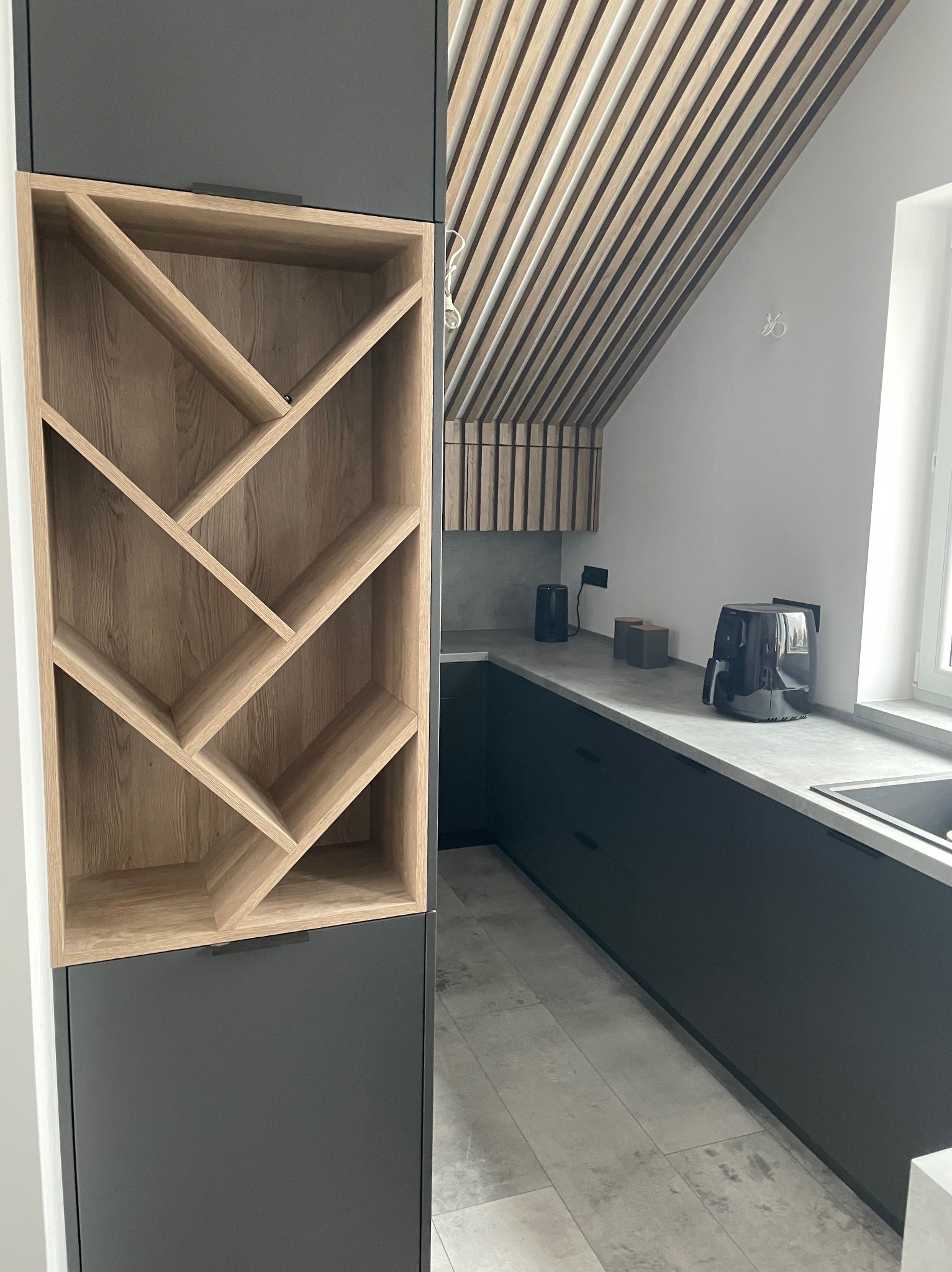 Nowoczesna kuchnia z szarymi szafkami i drewnianym regałem na wino. Uwagę przykuwa sufit z drewnianych listew, dodający wnętrzu ciepła i unikalnego charakteru.