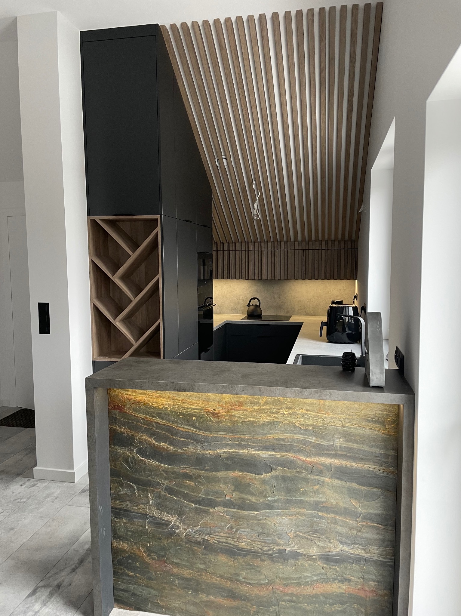 Nowoczesna kuchnia z czarnymi szafkami, drewnianym regałem na wino i dekoracyjnym panelem z kamienia naturalnego na wyspie kuchennej, oświetlona od dołu.