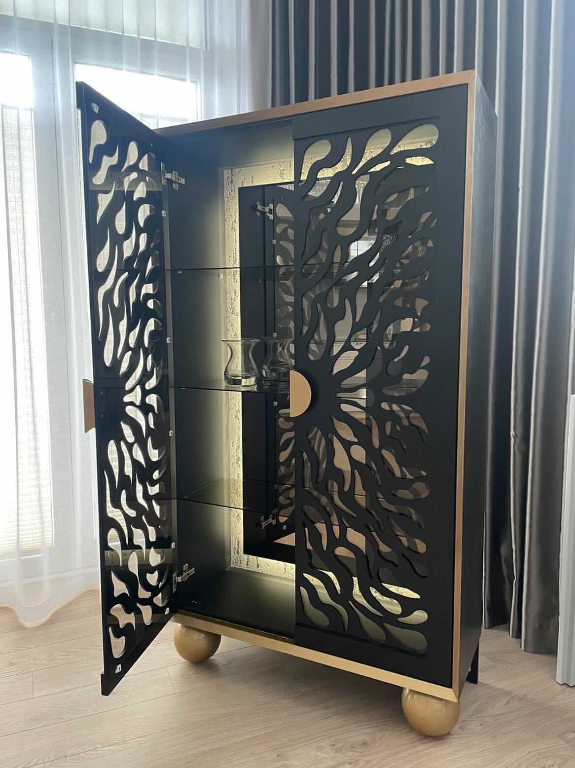 Czarna szafka z ażurowymi drzwiami, podświetlona od wewnątrz, z otwartymi drzwiami po lewej stronie, stojąca na drewnianej podłodze.