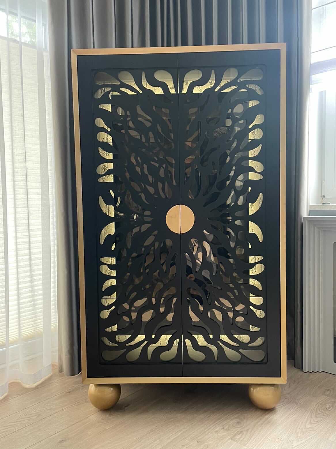 Czarna szafka z ażurowymi drzwiami i złotymi akcentami, stojąca na kulistych nóżkach, na tle okna z zasłonami. Dekoracyjny mebel w nowoczesnym wnętrzu.