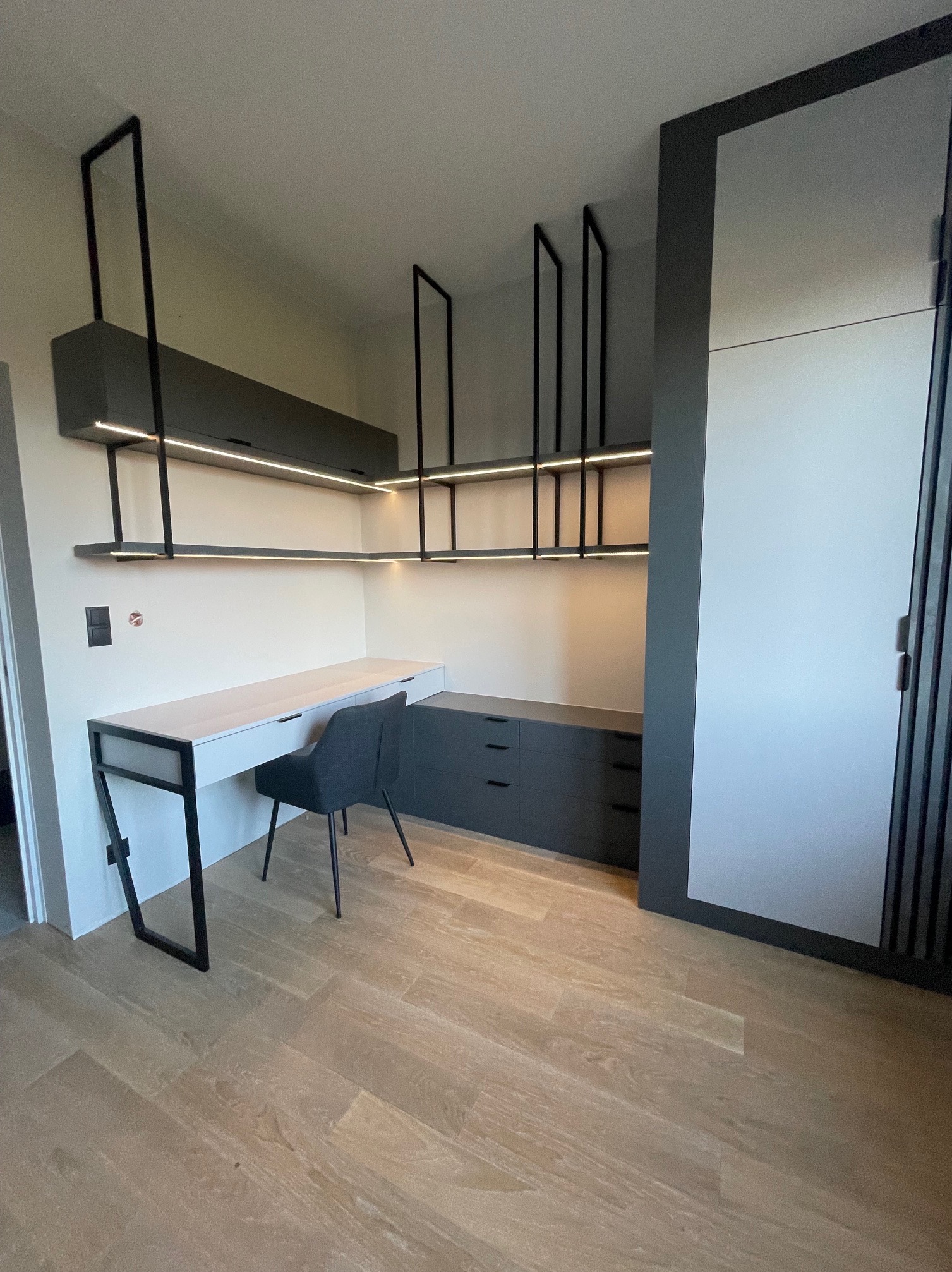 Nowoczesny pokój z biurkiem i szafą wnękową w odcieniach szarości, oświetlenie LED podkreśla minimalistyczny design. Widoczna drewniana podłoga i czarne metalowe elementy.