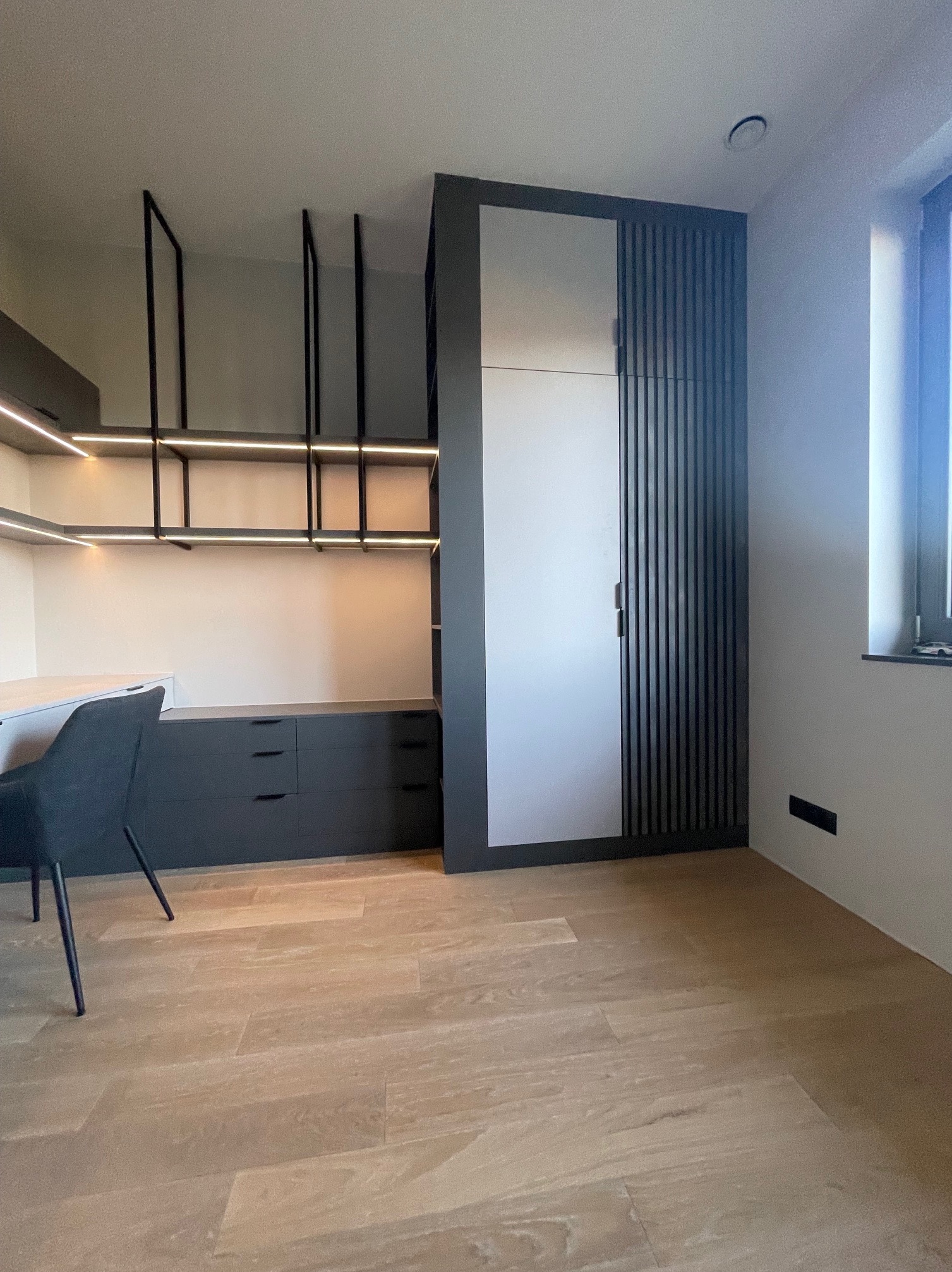 Nowoczesny pokój z biurkiem, szafą z lamelami i oświetleniem LED. Minimalistyczny design, drewniana podłoga, szare meble. Widok na okno z prawej strony.