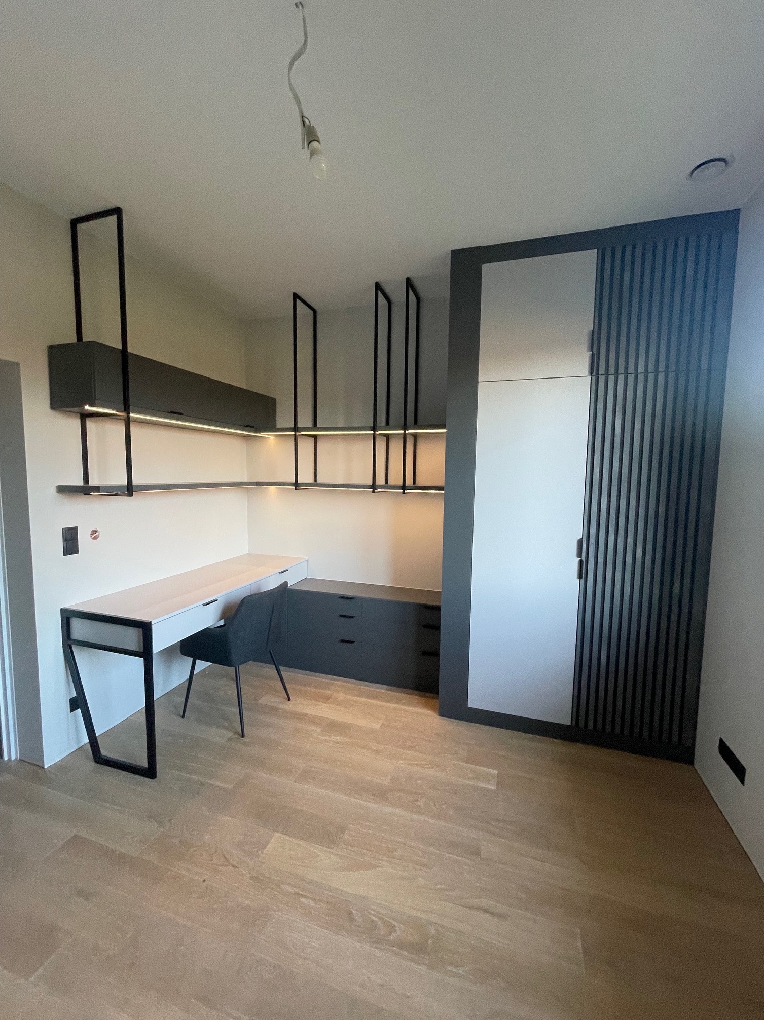 Nowoczesny pokój z biurkiem, szafą i półkami podświetlonymi LED. Minimalistyczny design w odcieniach szarości i bieli. Drewniana podłoga dodaje ciepła wnętrzu.