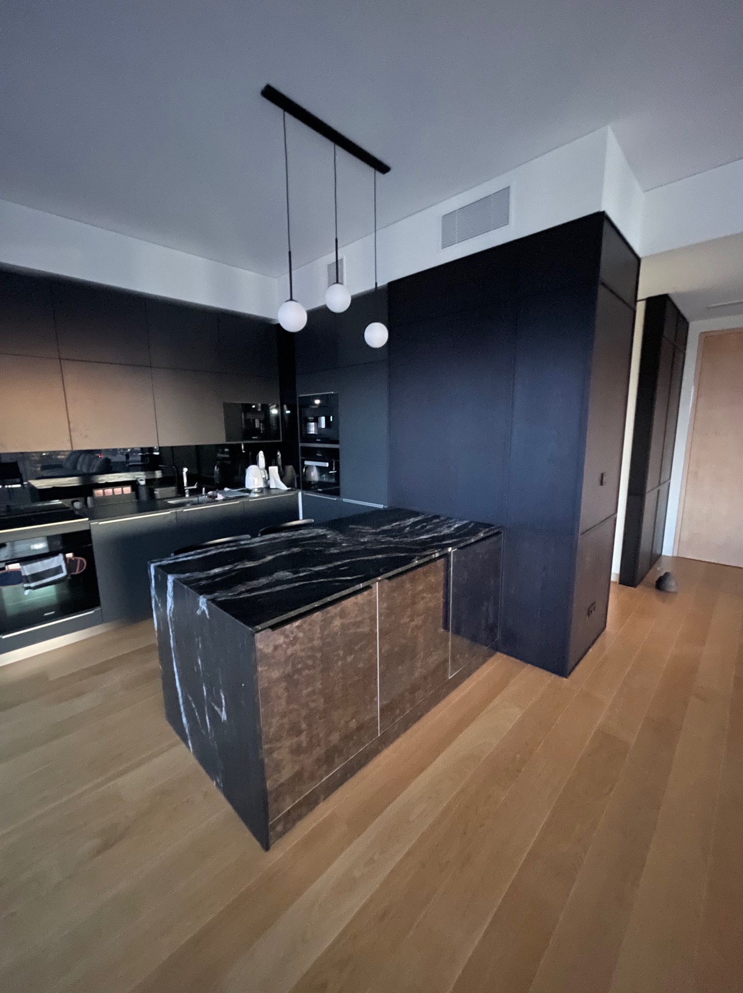 Nowoczesna kuchnia z czarnymi szafkami i wyspą z marmurowym blatem, oświetlona trzema kulistymi lampami wiszącymi. Parkiet na podłodze.