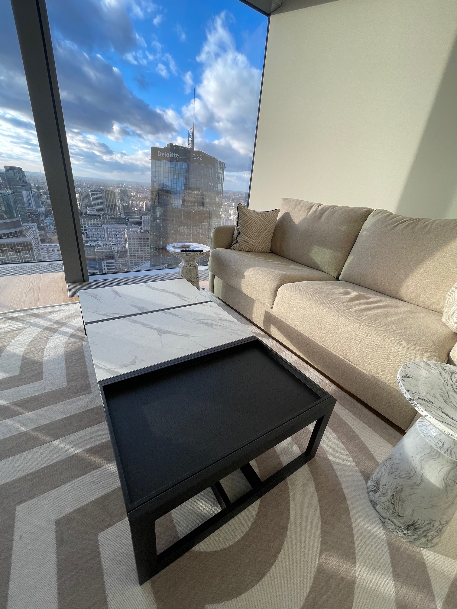 Nowoczesny salon z beżową sofą, stolikami kawowymi z marmurowymi blatami i widokiem na panoramę miasta przez okno. Minimalistyczny design, jasne wnętrze.