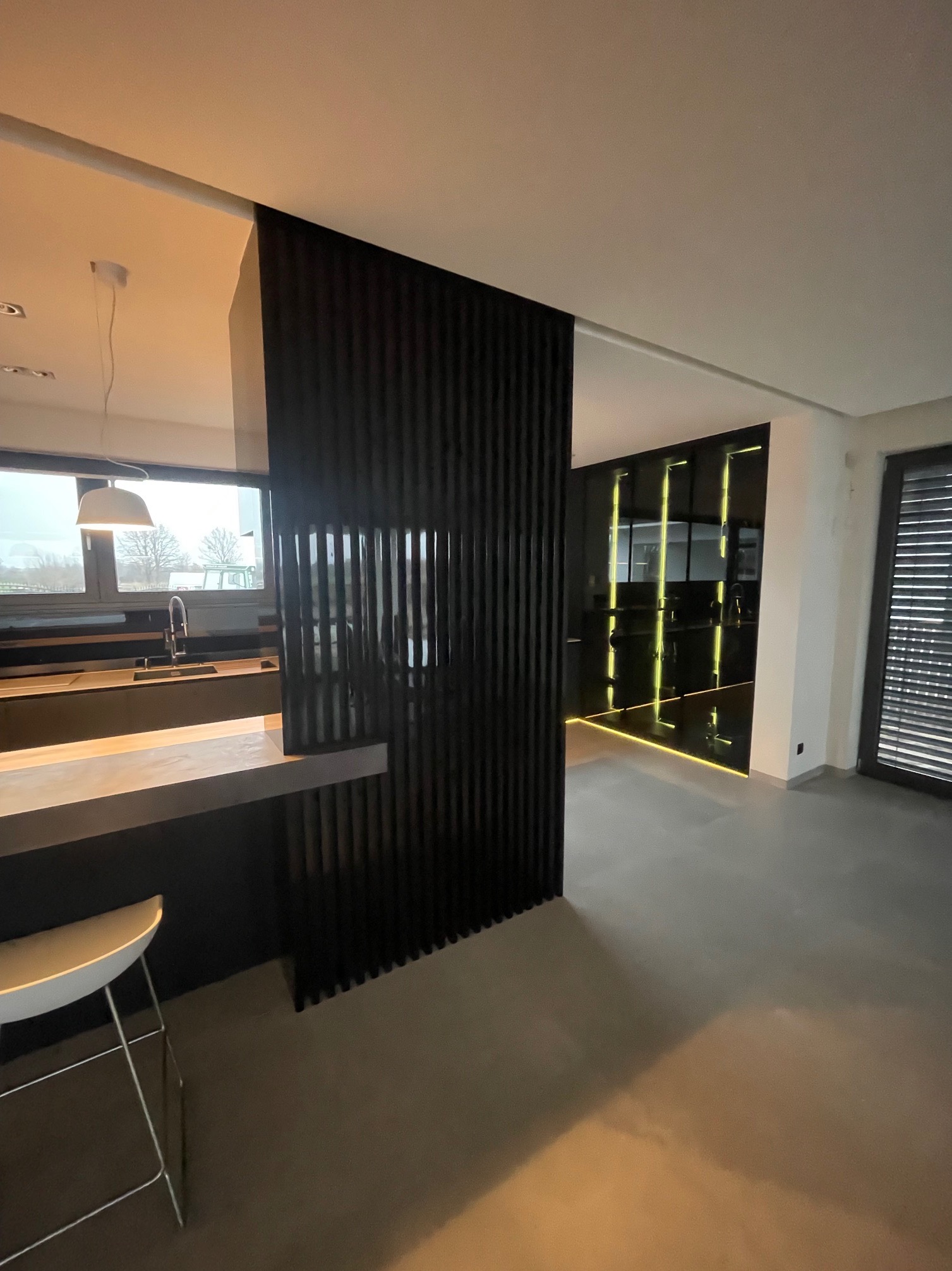 Nowoczesna kuchnia z czarną, lamelową ścianką działową i wyspą. W tle szafa wnękowa z podświetleniem LED. Minimalistyczny design, szara podłoga.