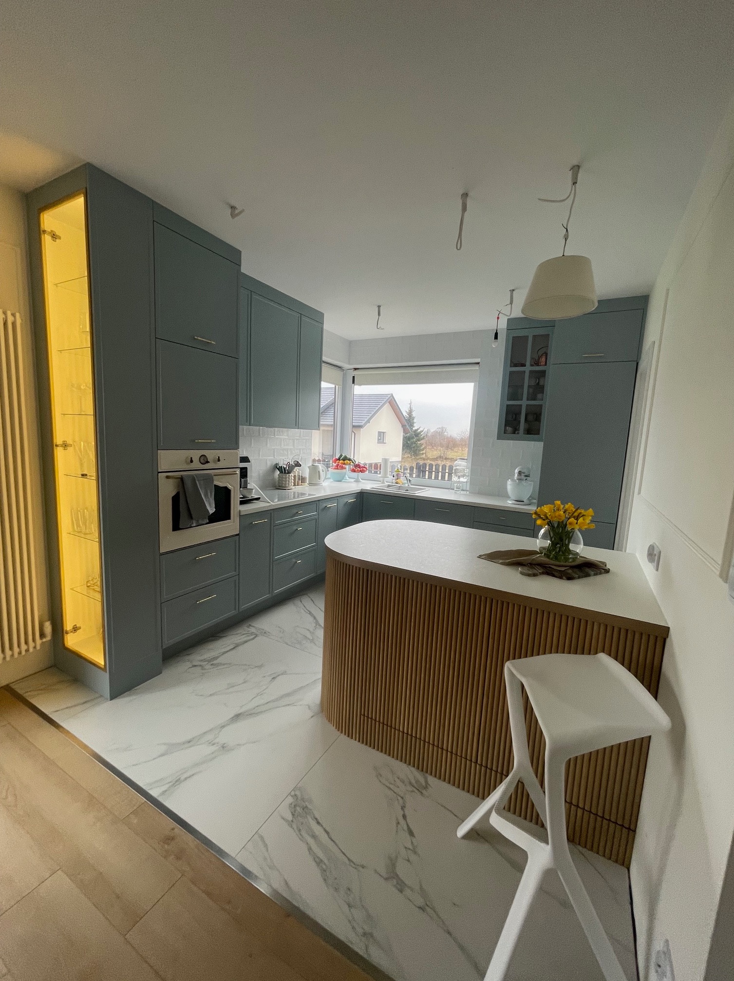 Niebieska kuchnia z wyspą o zaokrąglonym froncie z drewnianych listewek, biały blat i krzesło barowe. Na blacie żółte kwiaty w wazonie. Nowoczesny design.