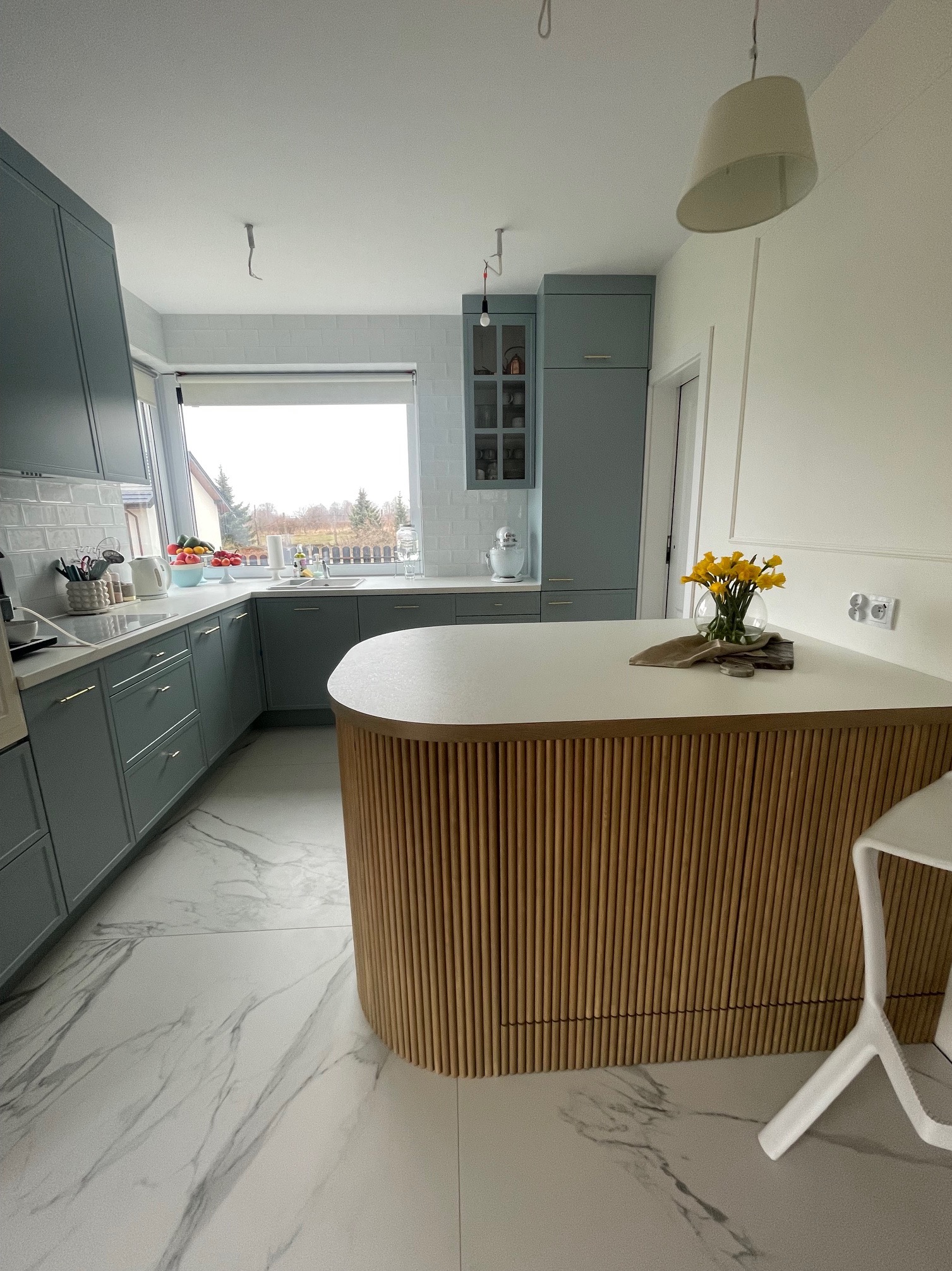 Nowoczesna kuchnia w odcieniach szarości z drewnianą wyspą i marmurową podłogą. Na blacie wyspy stoi wazon z żółtymi kwiatami, dodając akcentu kolorystycznego.