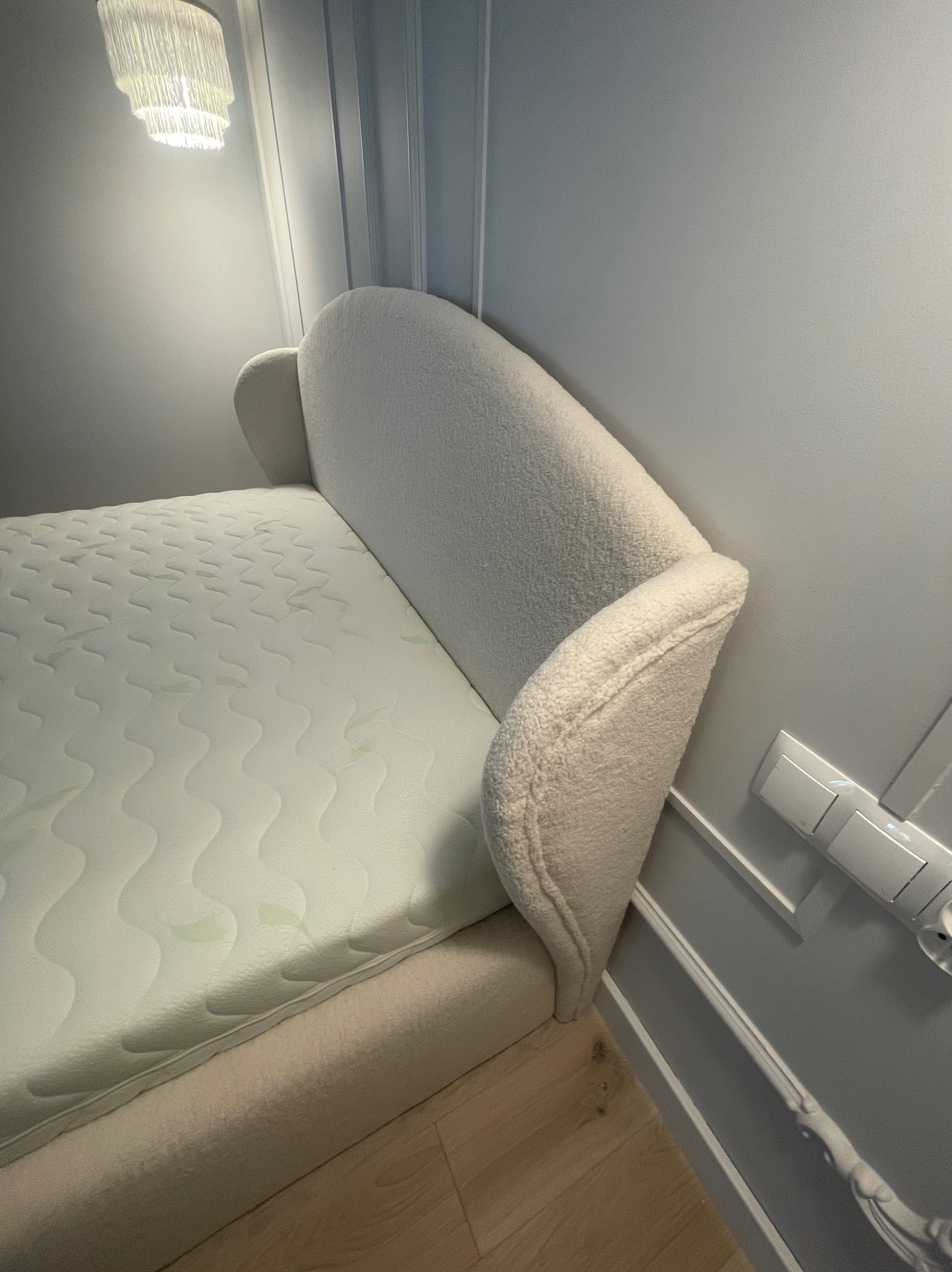 Beżowe łóżko z wełnianym zagłówkiem w minimalistycznej sypialni. Widoczny materac i fragment podłogi. Nad łóżkiem lampa. Neutralna kolorystyka.