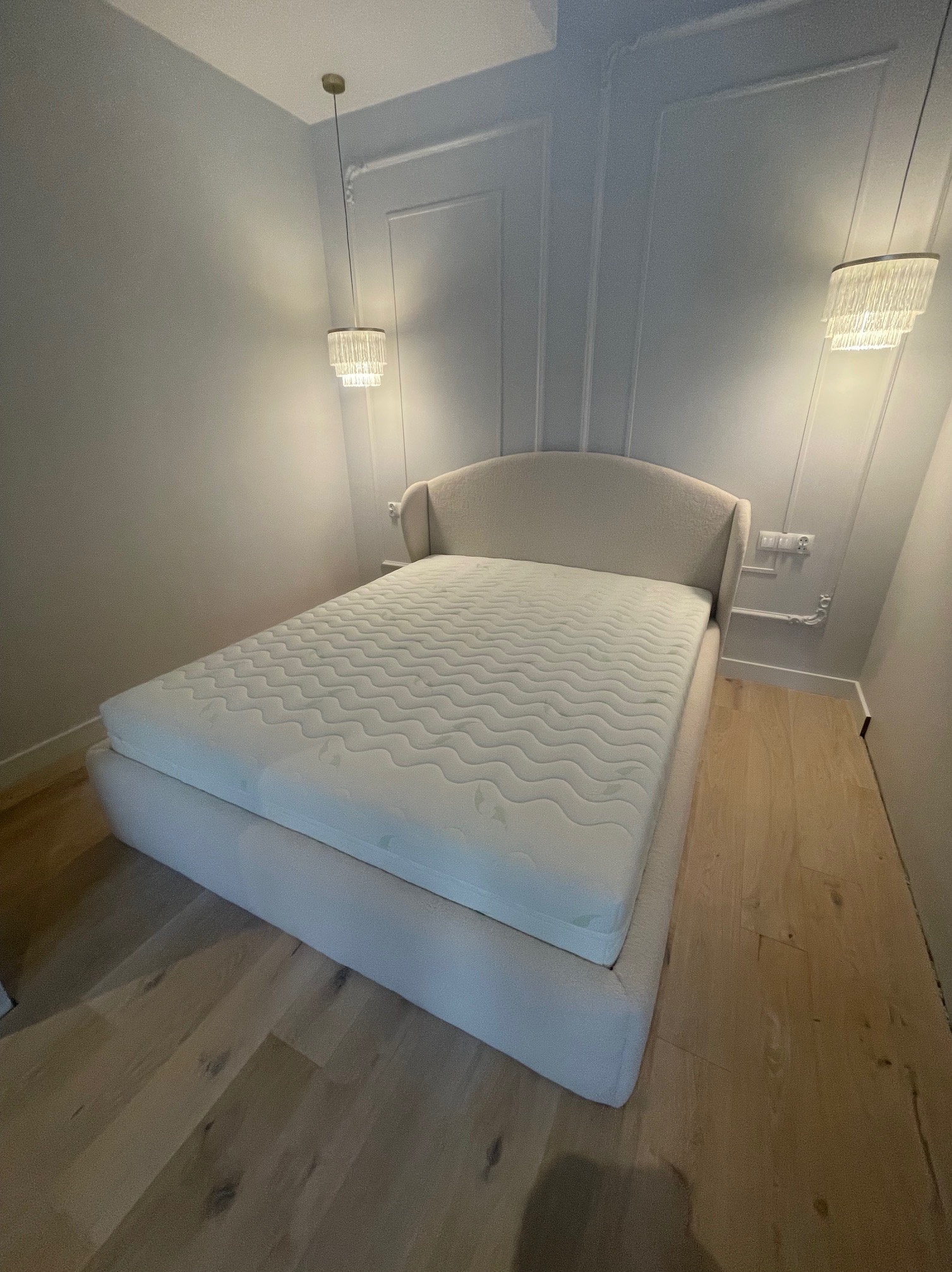 Eleganckie łóżko tapicerowane w jasnej tonacji, z wysokim wezgłowiem i materacem, w minimalistycznej sypialni z drewnianą podłogą i dekoracyjnymi lampami wiszącymi.