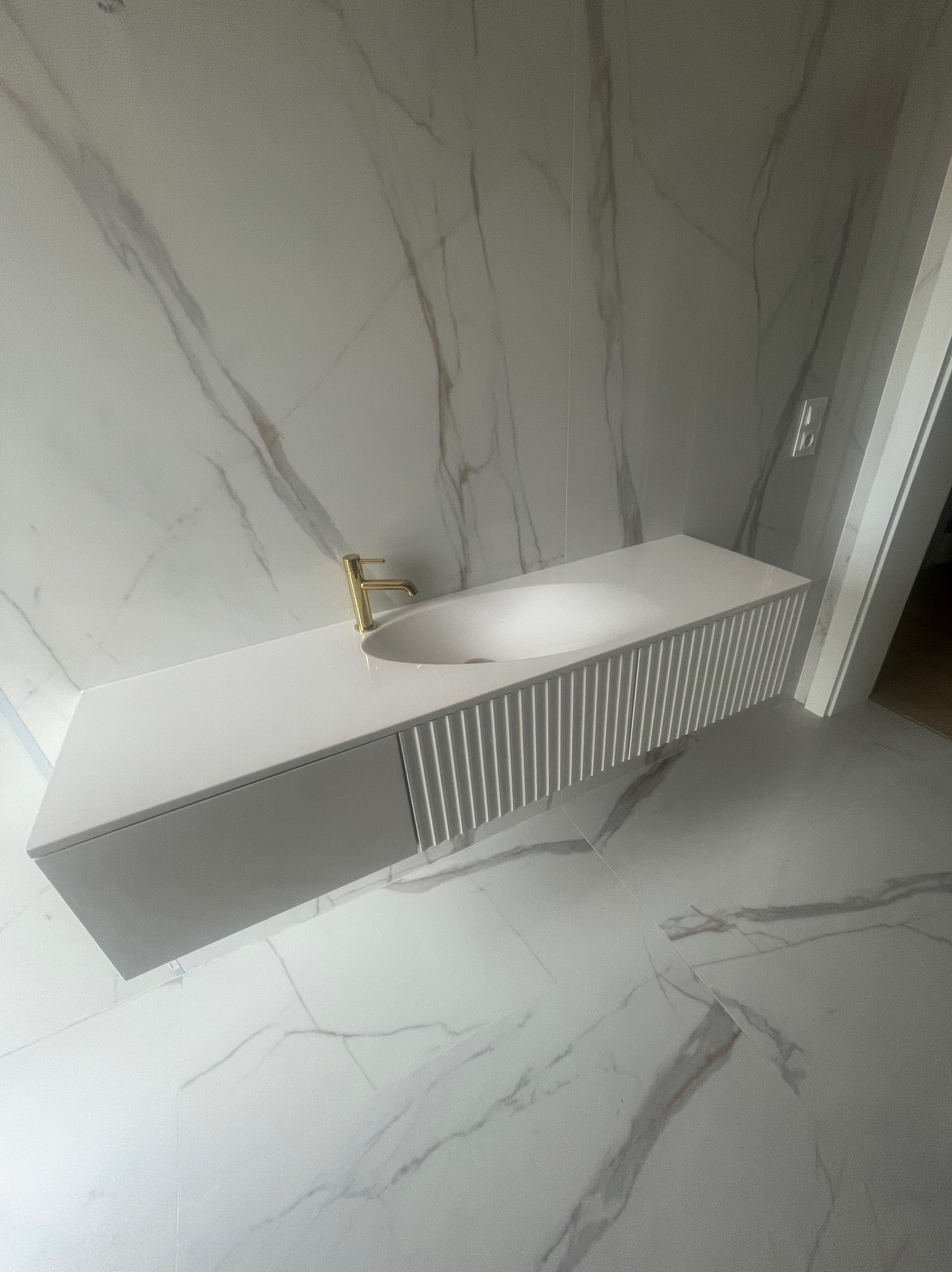 Biała, podwieszana szafka łazienkowa z umywalką i złotą baterią na tle marmurowych płytek. Nowoczesny design z frezowanymi frontami.