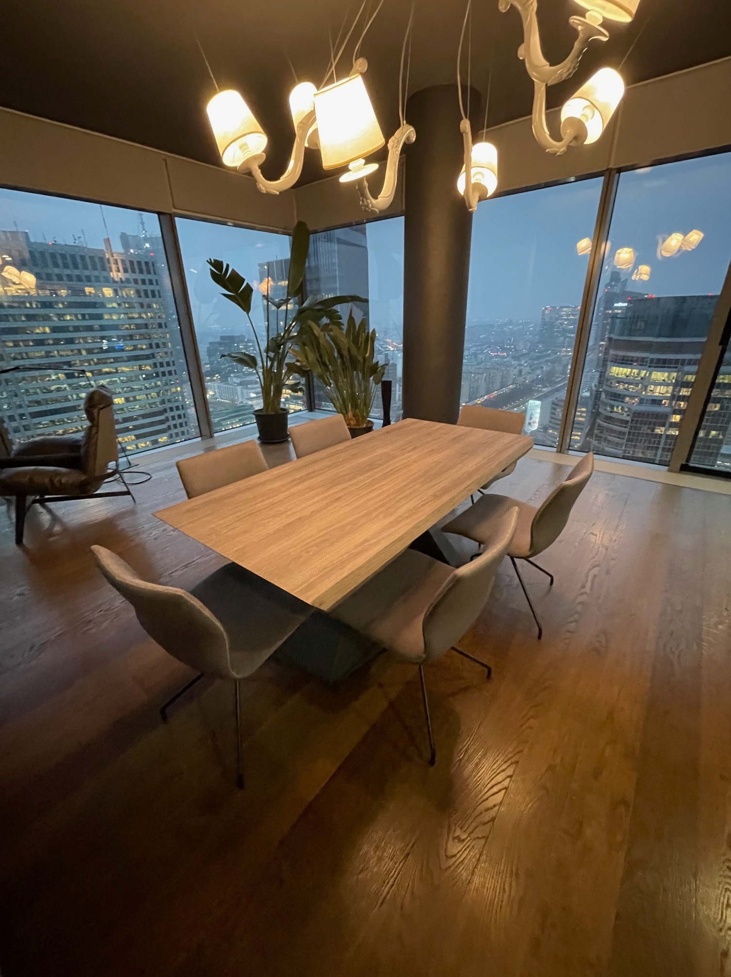 Nowoczesna jadalnia z drewnianym stołem i krzesłami na tle panoramy miasta za oknem. Designerska lampa sufitowa i drewniana podłoga dodają elegancji.