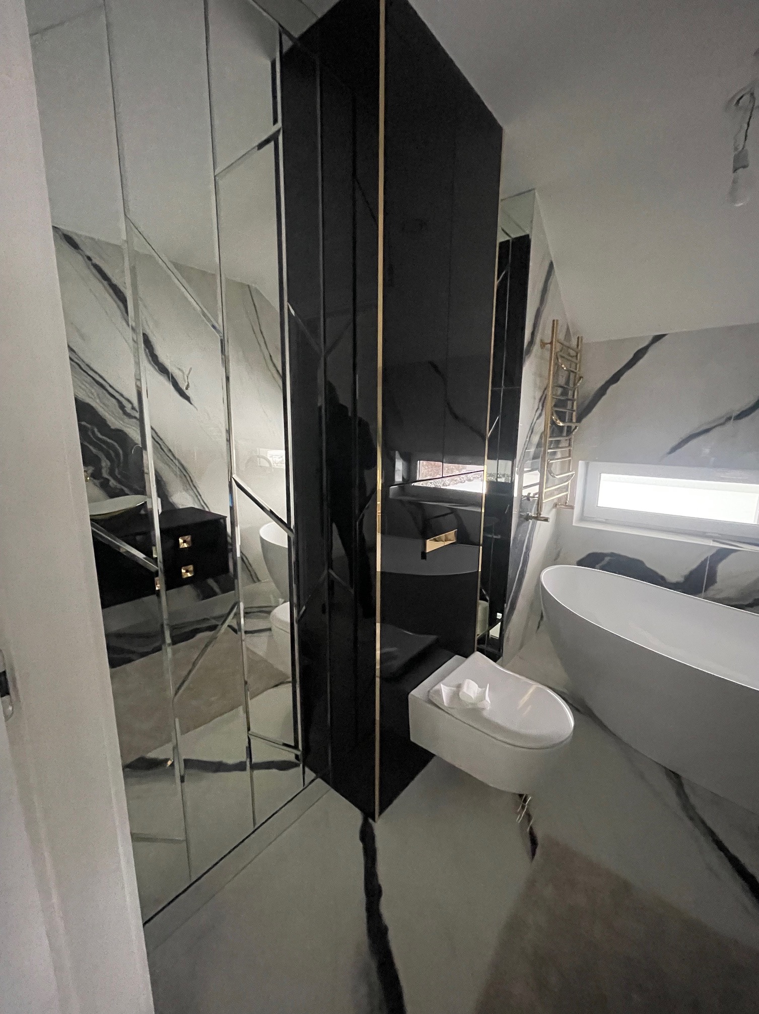 Nowoczesna łazienka z czarną szafą w lustrzanych panelach, białą wanną wolnostojącą i marmurowymi płytkami. Widok z perspektywy wejścia, akcent na geometrię i odbicia.
