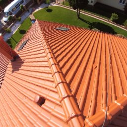 Malowanie Dachów - Czerwony dach z dachówki ceramicznej, widok z góry, z widocznymi oknami dachowymi i rynną. W tle trawnik i budynki. Ujęcie z bliska, podkreślające strukturę dachówki.