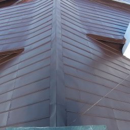 Malowanie Dachów - Brązowy dach z blachy trapezowej, widok z góry, z kominem w kolorze białym z zielonym daszkiem. Widoczne obróbki blacharskie i fragmenty zabudowy.