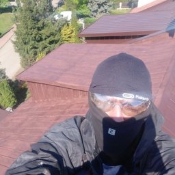 Malowanie Dachów - Pracownik w stroju ochronnym na brązowym dachu z blachy trapezowej, widok z góry na okoliczne domy i drzewa w słoneczny dzień.