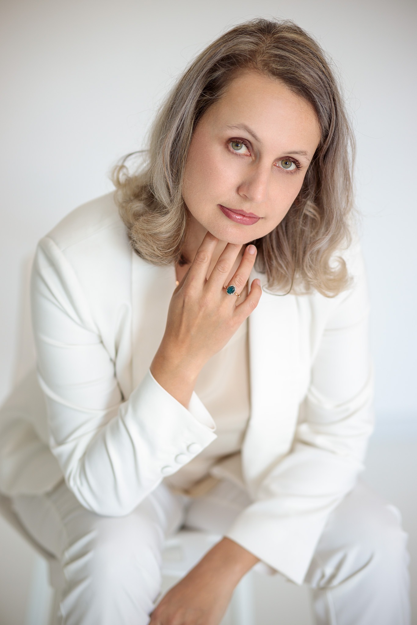 Portret kobiety w białym garniturze na jasnym tle, z dłonią przy szyi i pierścionkiem z zielonym kamieniem. Profesjonalny wizerunek.