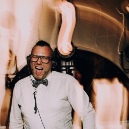 DJ Głośny - Maciej Głos - Uśmiechnięty DJ w muszce i okularach, z słuchawkami na uszach, w trakcie występu, z oświetleniem scenicznym w tle. Dynamiczne ujęcie.