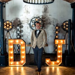 DJ Głośny - Maciej Głos - Uśmiechnięty DJ w beżowej marynarce i muszce, stoi między podświetlanymi literami 'DJ' na tle dekoracji scenicznej z reflektorami i rysunkiem koni.