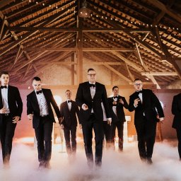 DJ Głośny - Maciej Głos - Eleganccy mężczyźni w smokingach idą w dymie na tle drewnianej konstrukcji z oświetleniem, tworząc sceniczny efekt. Ujęcie z imprezy.