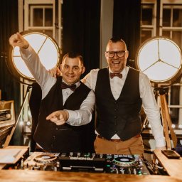 DJ Głośny - Maciej Głos - Uśmiechnięci DJ-e w muszkach, za konsoletą Denon DJ, wskazują palcami, w tle lampy reflektorowe i okno. Profesjonalne nagłośnienie i oświetlenie imprezy.