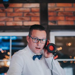 DJ Głośny - Maciej Głos - DJ w okularach i muszce, z słuchawkami na szyi, w trakcie występu na tle ceglanej ściany i okna z rozmytymi światłami. Profesjonalne nagłośnienie imprezy.