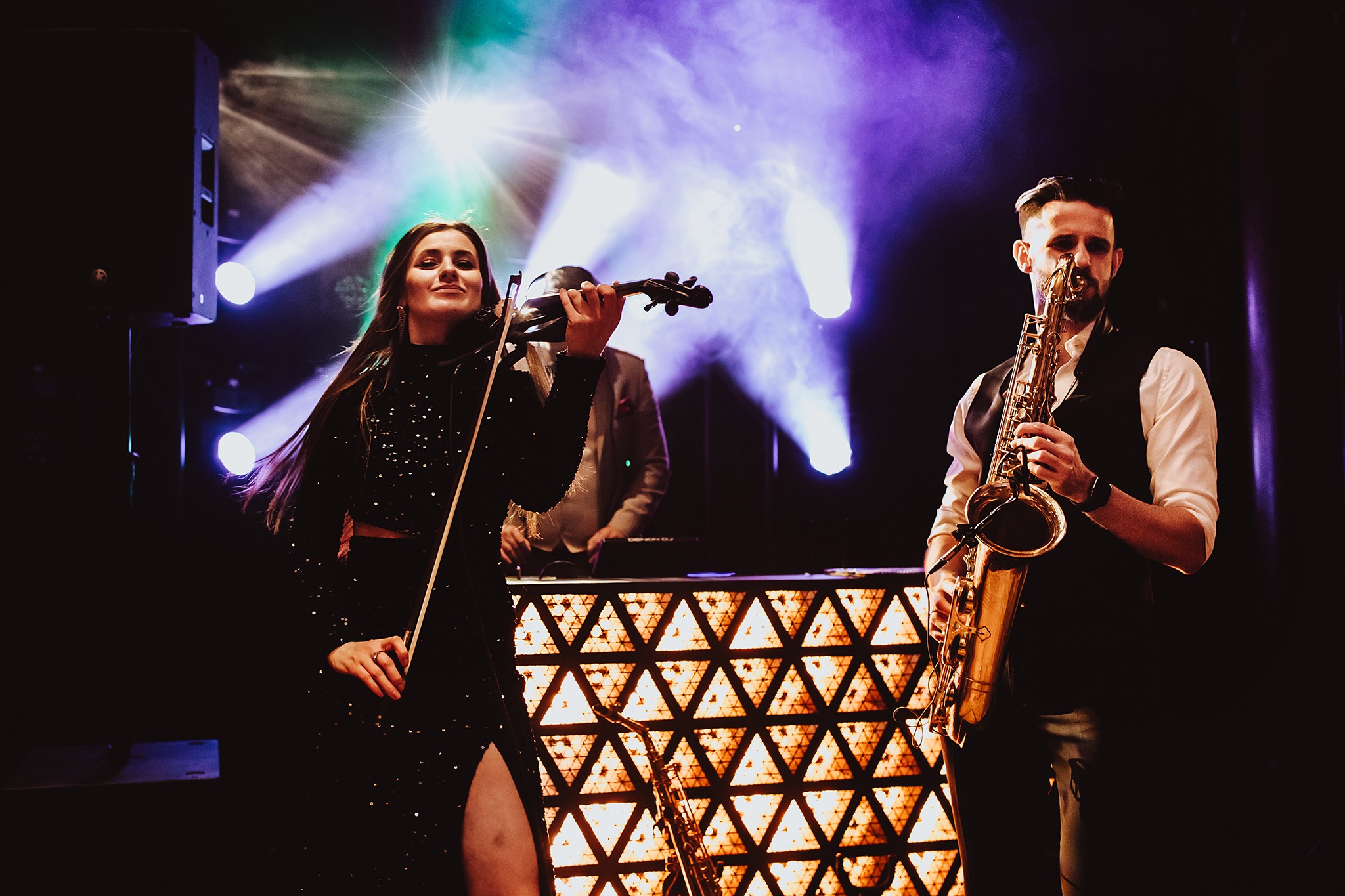 Zespół muzyczny na scenie: skrzypaczka w błyszczącej sukience i saksofonista z brodą, oświetleni reflektorami, z DJ-em w tle. Dynamiczne ujęcie koncertowe.