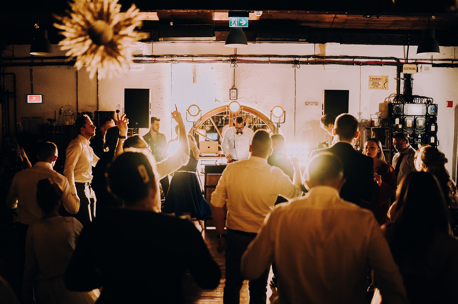 DJ w muszce gra muzykę na imprezie, otoczony tańczącymi ludźmi. W tle industrialne elementy dekoracyjne i ciepłe oświetlenie.