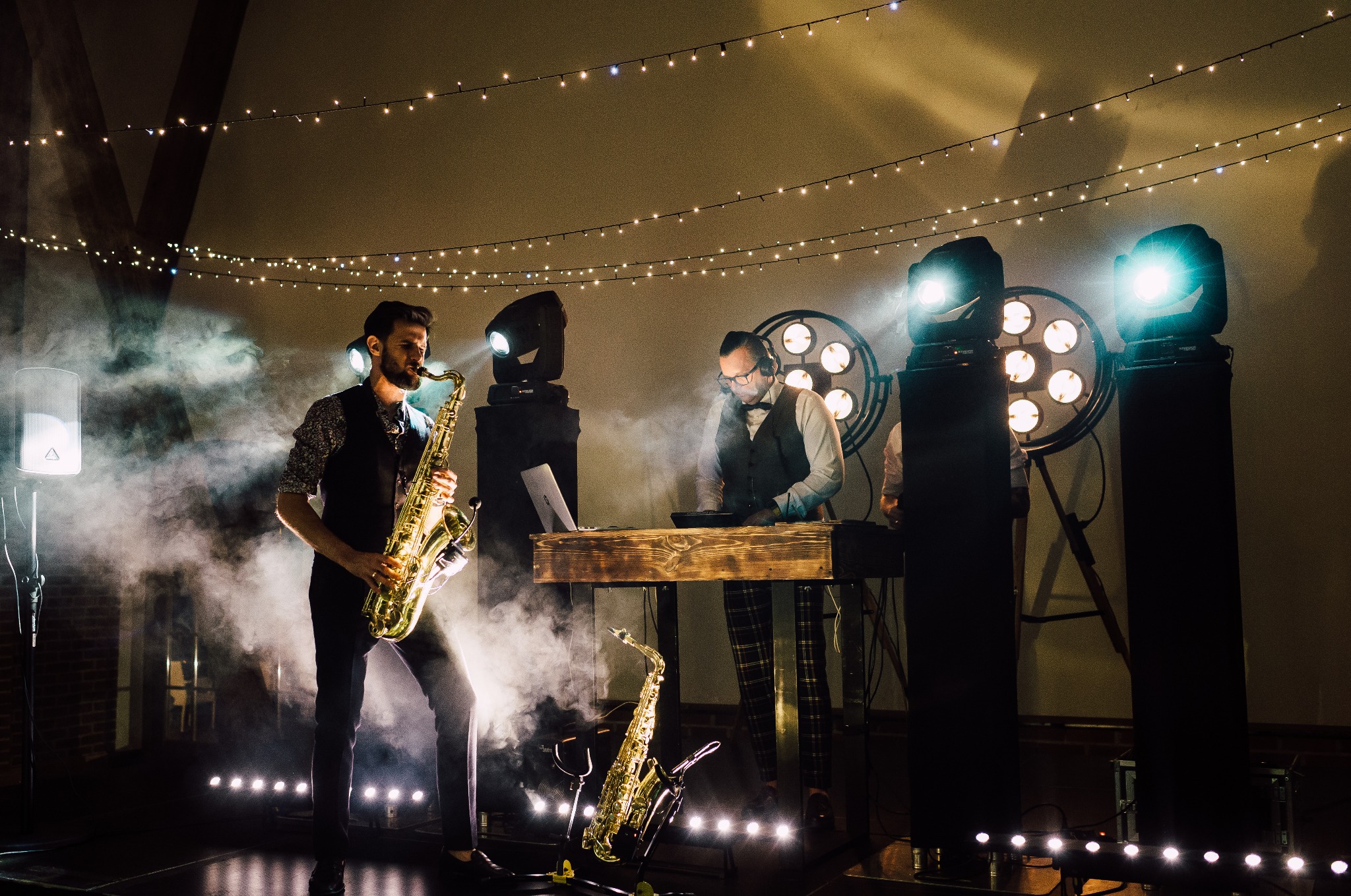 Saksofonista w akcji, DJ przy drewnianym stanowisku, profesjonalne oświetlenie sceniczne i dekoracyjne lampki tworzą wyjątkowy klimat podczas występu.