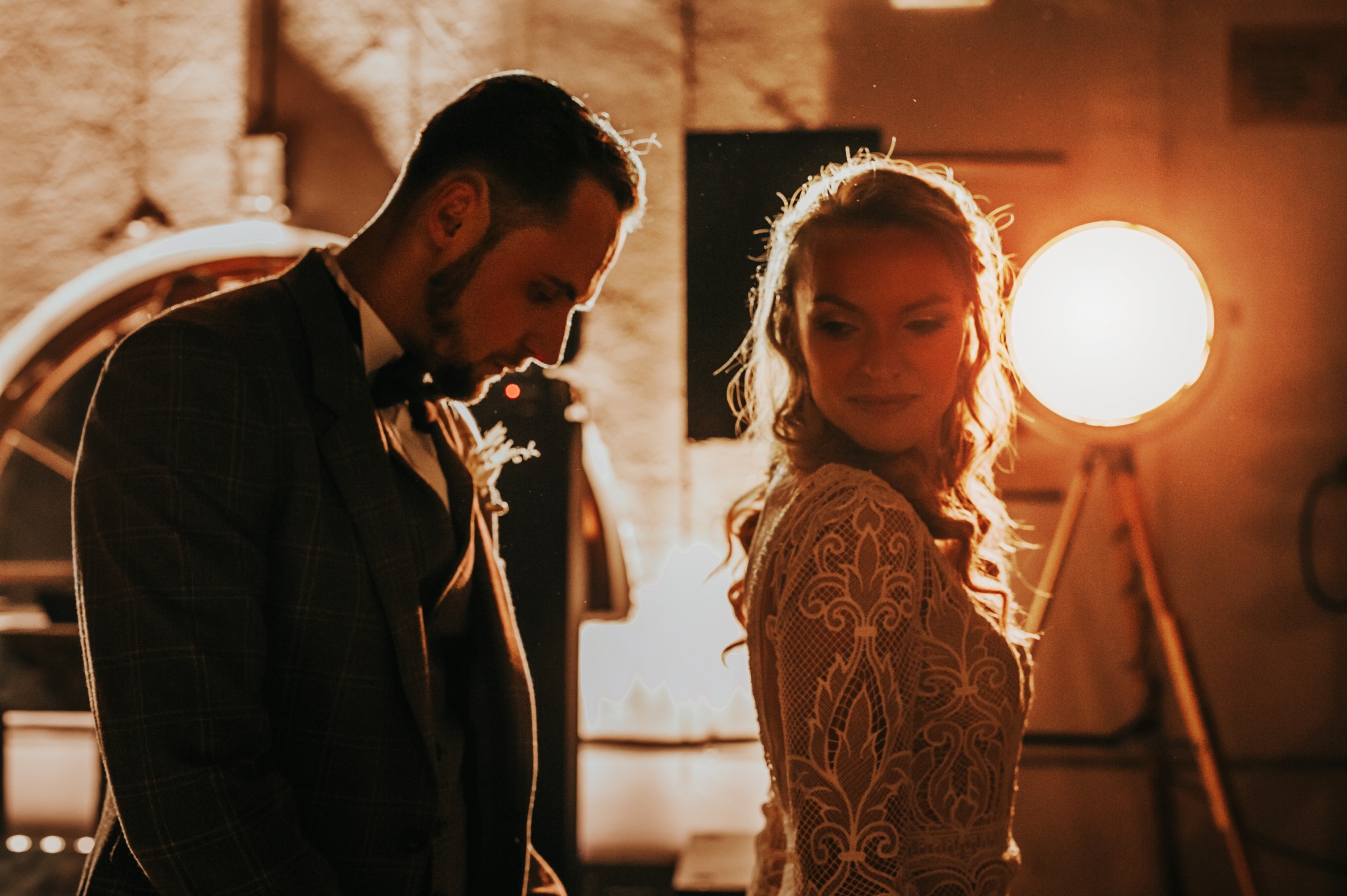 Para młoda w ciepłym oświetleniu studyjnym: on w garniturze w kratę, ona w koronkowej sukni, oboje zamyśleni, z lampą studyjną w tle. Romantyczna sesja.