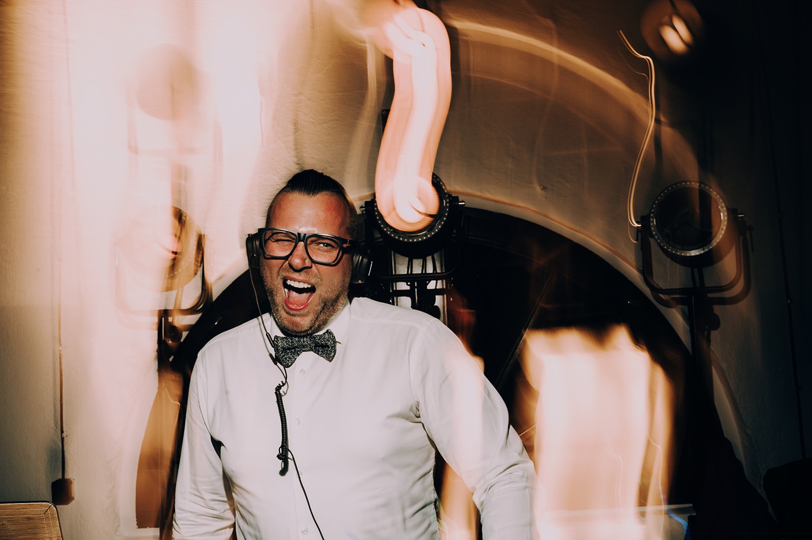 Uśmiechnięty DJ w muszce i okularach, z słuchawkami na uszach, w trakcie występu, z oświetleniem scenicznym w tle. Dynamiczne ujęcie.