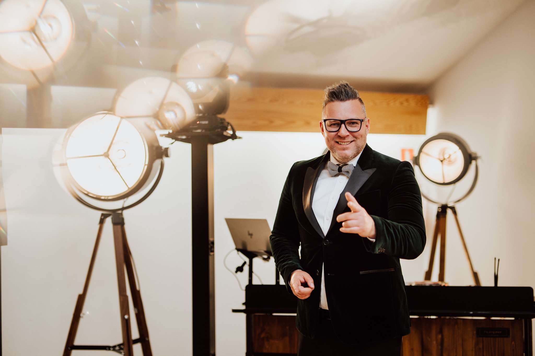 Uśmiechnięty mężczyzna w aksamitnej marynarce i muszce, wskazujący palcem, na tle lamp studyjnych i laptopa. Scena sugeruje przygotowania do występu lub eventu.