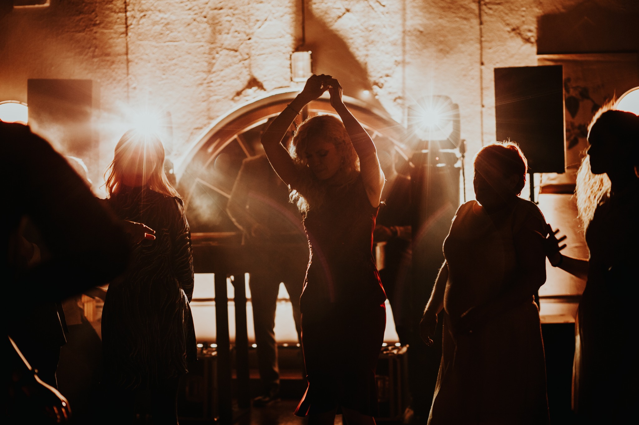 Sylwetki tańczących osób w ciepłym oświetleniu na tle sceny z elementami dekoracyjnymi. Dynamiczna atmosfera imprezy z rozmytymi postaciami w ruchu.