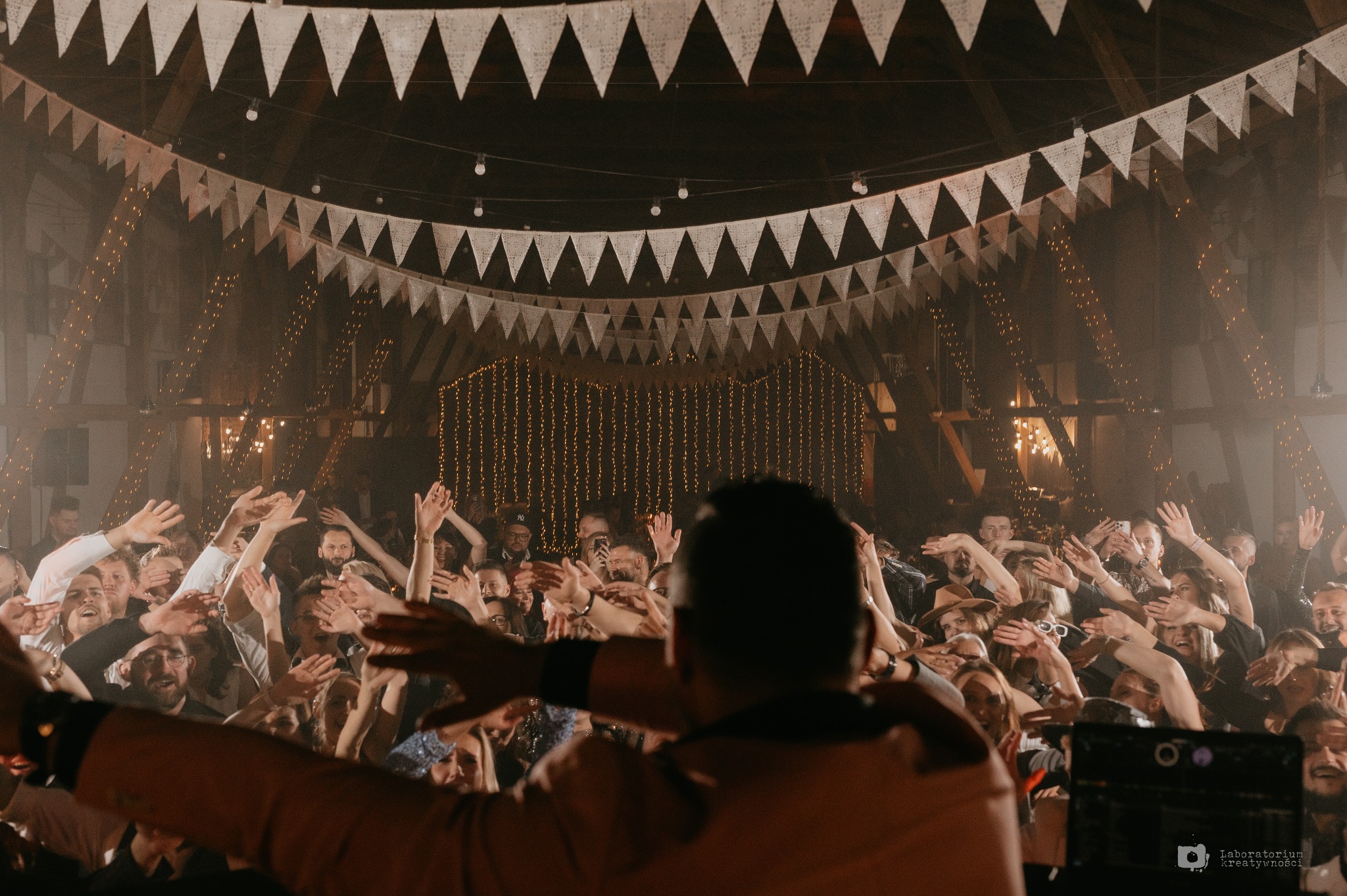 Rozbawiony tłum z podniesionymi rękami podczas koncertu w rustykalnej sali z dekoracjami z girland i światełek, widziany zza pleców DJ-a.