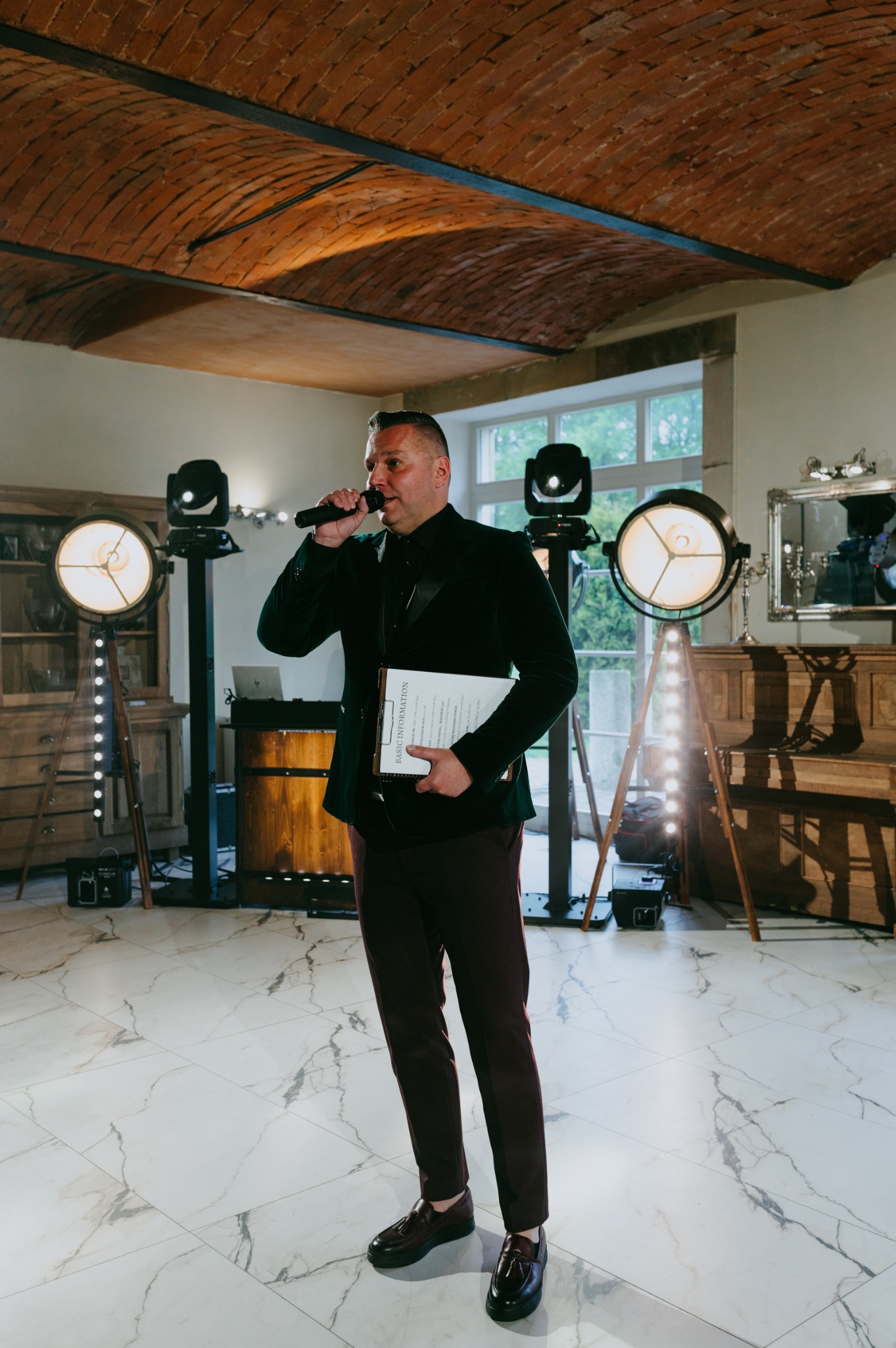 Prezenter w zielonym aksamitnym smokingu, z mikrofonem i kartką, na tle sceny z oświetleniem, w eleganckim wnętrzu z ceglanym sklepieniem.