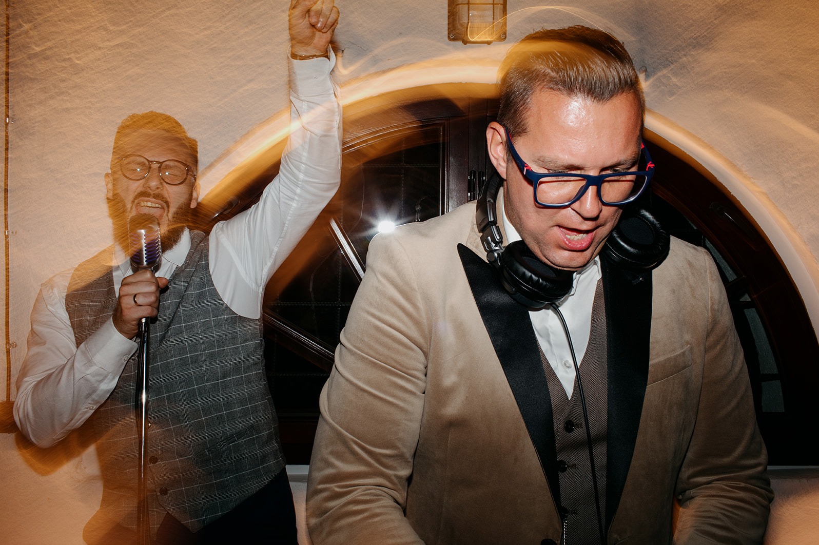 Dwóch mężczyzn w eleganckich strojach, jeden śpiewa do mikrofonu z uniesioną ręką, drugi z słuchawkami na szyi, w tle rozmyte światła.