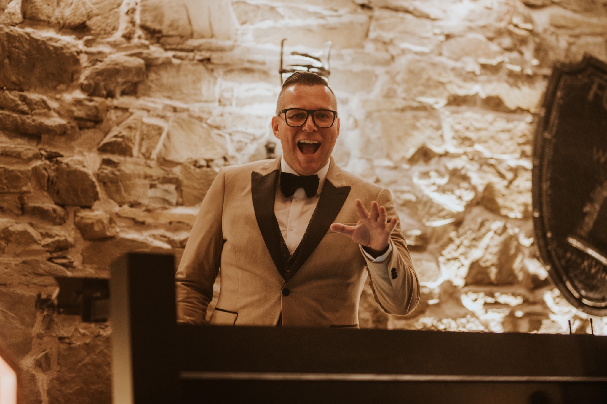 Uśmiechnięty mężczyzna w beżowym smokingu i muszce, za stołem DJ-a, na tle kamiennej ściany i dekoracji w stylu rustykalnym, z otwartymi ustami i uniesioną dłonią.