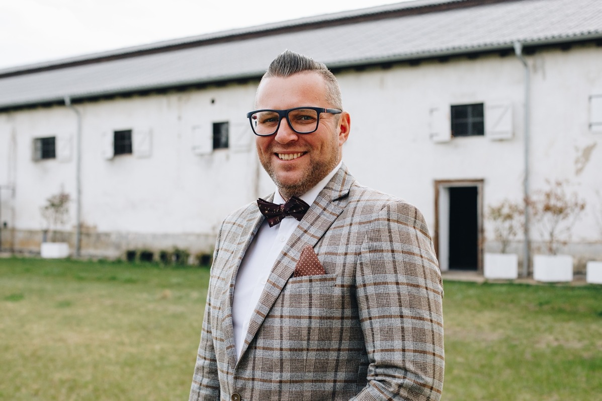 Uśmiechnięty mężczyzna w kraciastej marynarce i muszce, na tle białego budynku z oknami. Elegancki strój, plener, jasne oświetlenie.