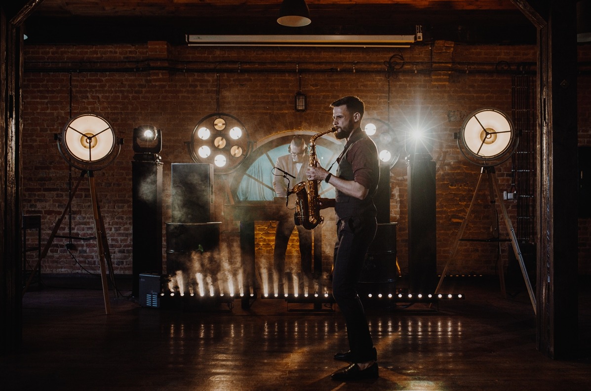 Saksofonista w akcji na tle ceglanej ściany i oświetlenia scenicznego. Drugi muzyk w tle. Klimatyczne światło i dym tworzą unikalny nastrój.