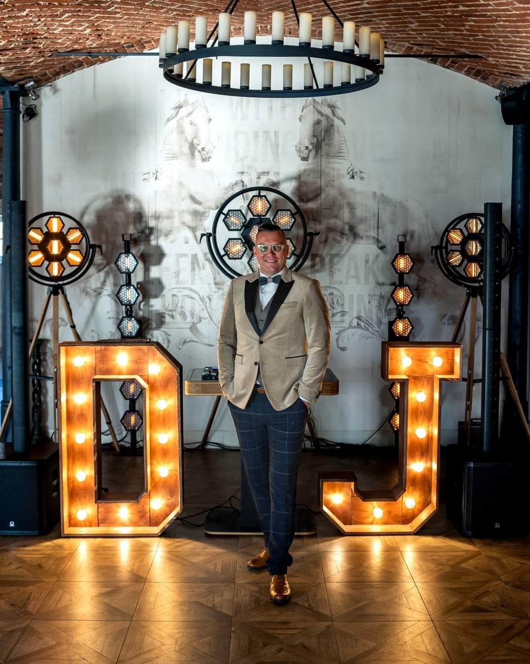 Uśmiechnięty DJ w beżowej marynarce i muszce, stoi między podświetlanymi literami 'DJ' na tle dekoracji scenicznej z reflektorami i rysunkiem koni.