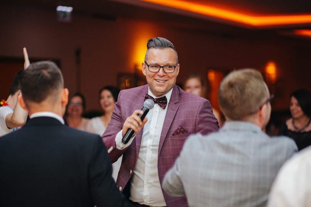 Uśmiechnięty konferansjer w bordowym garniturze w kratę, z mikrofonem na tle rozbawionych gości weselnych i ciepłego oświetlenia sali.
