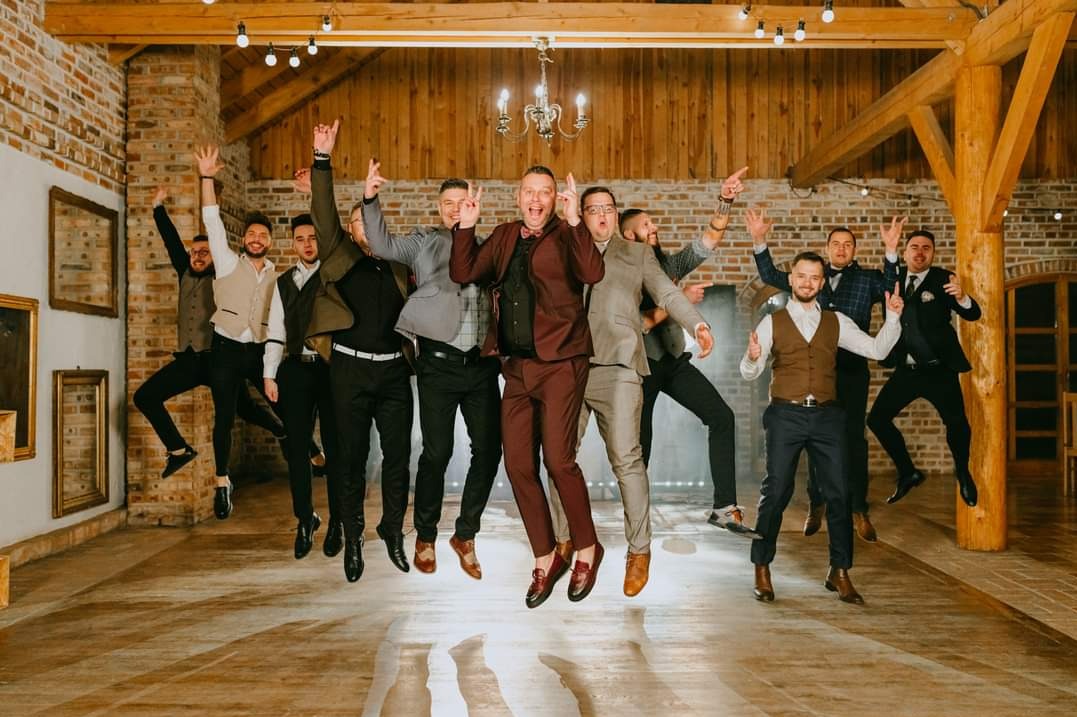 Rozbawiona grupa mężczyzn w garniturach skacze radośnie w przestronnym wnętrzu z ceglanymi ścianami i drewnianymi belkami, świętując udane wydarzenie.