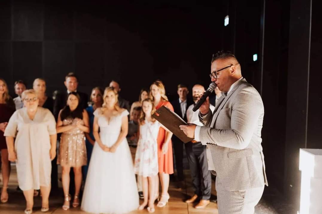 Konferansjer w szarym garniturze prowadzi uroczystość ślubną, trzymając mikrofon i notatki. W tle para młoda i goście.