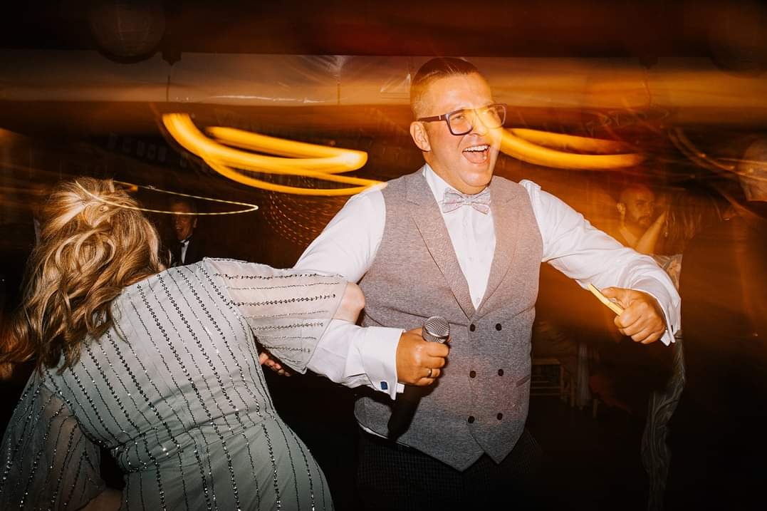 Uśmiechnięty mężczyzna w okularach i kamizelce śpiewa do mikrofonu podczas imprezy, w tle tańcząca kobieta i rozmyte światła.