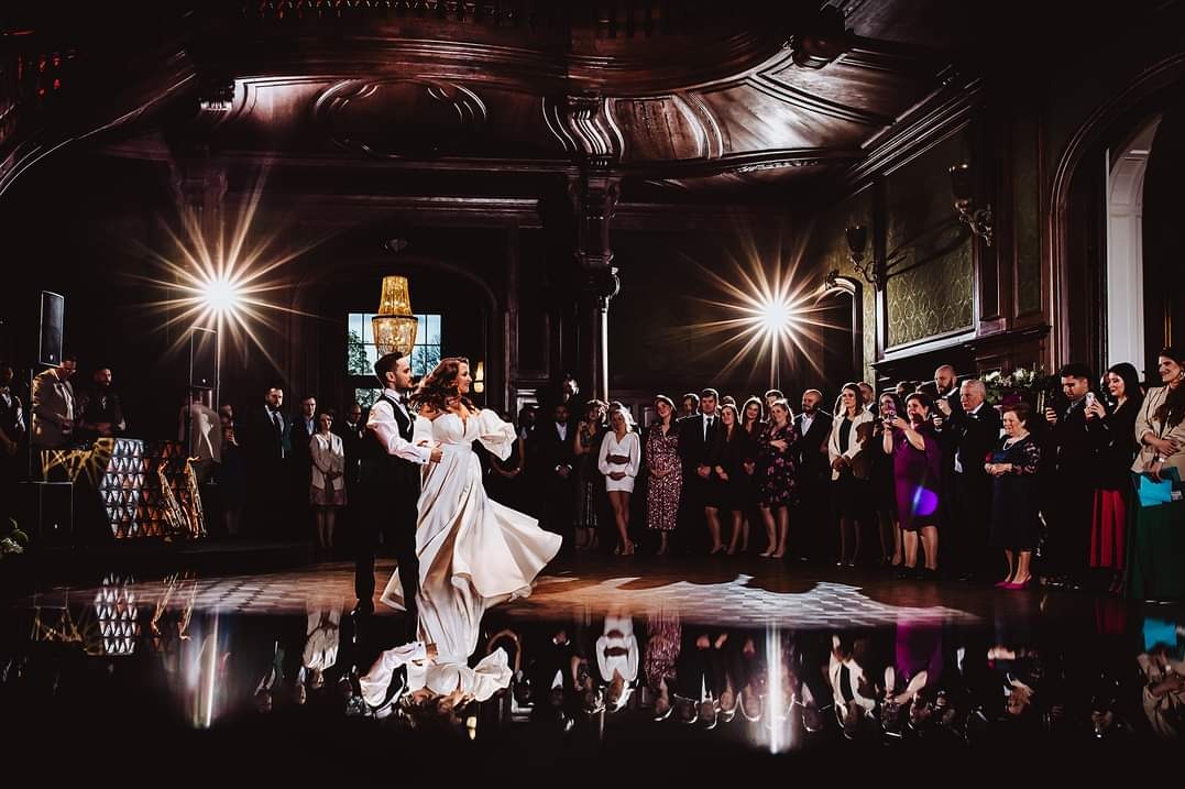 Para młoda tańczy pierwszy taniec na parkiecie z lustrzanym odbiciem, otoczona gośćmi w eleganckiej sali z dekoracyjnym sufitem i oświetleniem.