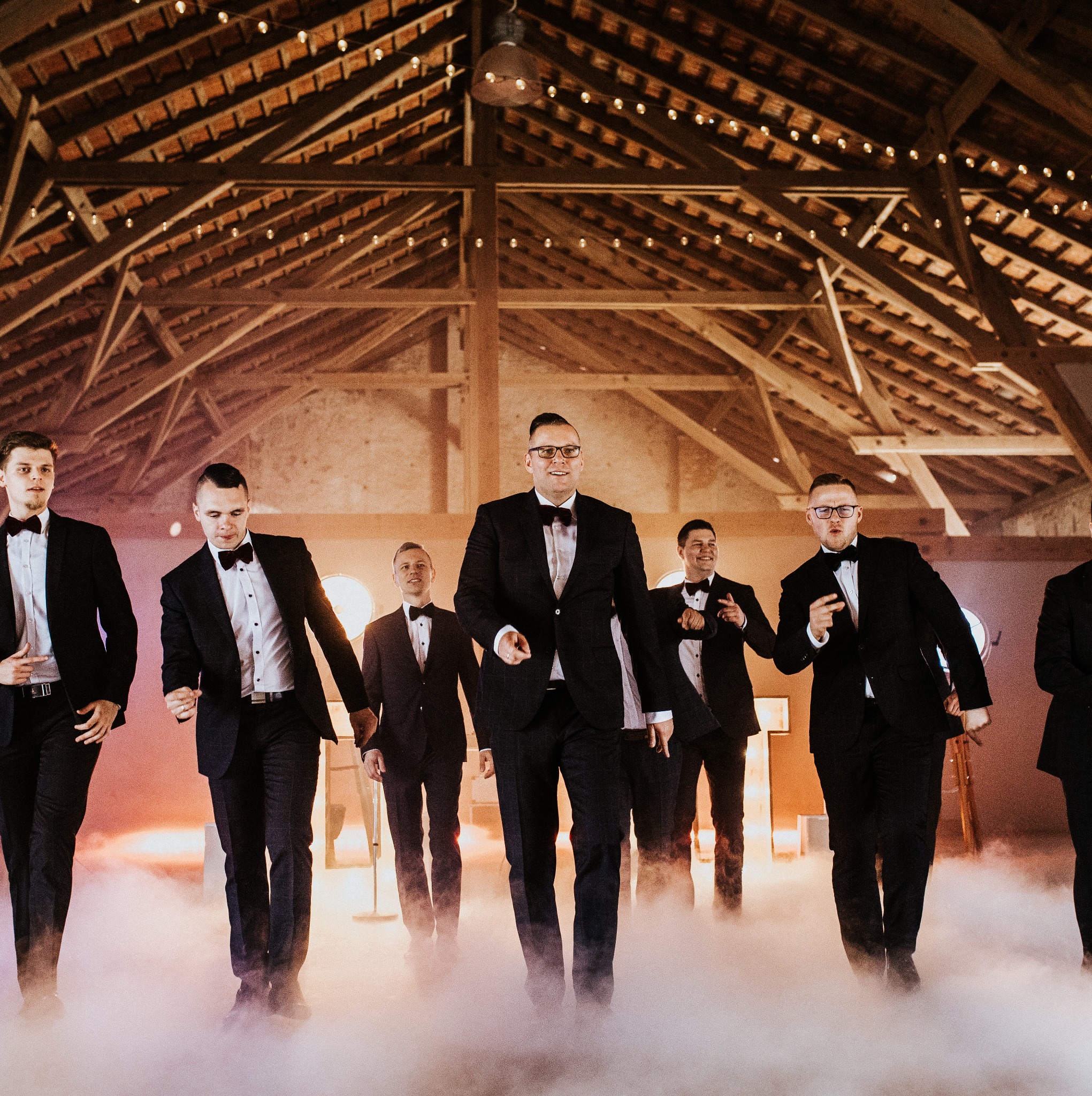 Eleganccy mężczyźni w smokingach idą w dymie na tle drewnianej konstrukcji z oświetleniem, tworząc sceniczny efekt. Ujęcie z imprezy.