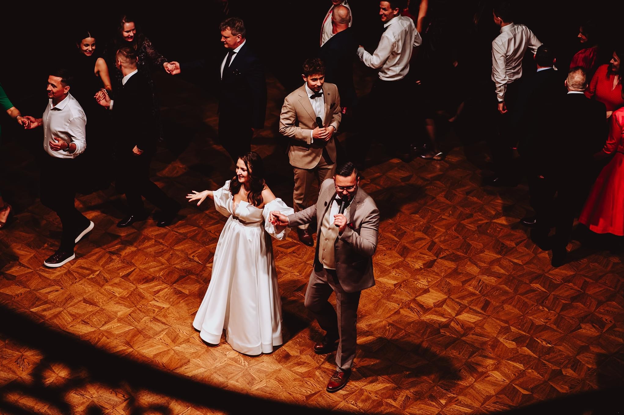 Zespół muzyczny na weselu w trakcie występu. Wokalistka w białej sukni i wokalista w szarym garniturze śpiewają na parkiecie z parkietu w otoczeniu gości.